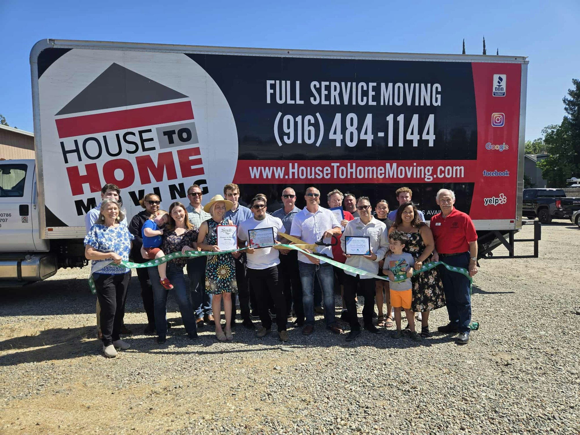 A group of people gathers for a ribbon-cutting ceremony in front of a "House to Home Moving" truck.