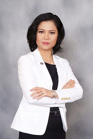 A woman in a white blazer and black attire, with crossed arms, against a grey background. Professional portrait.