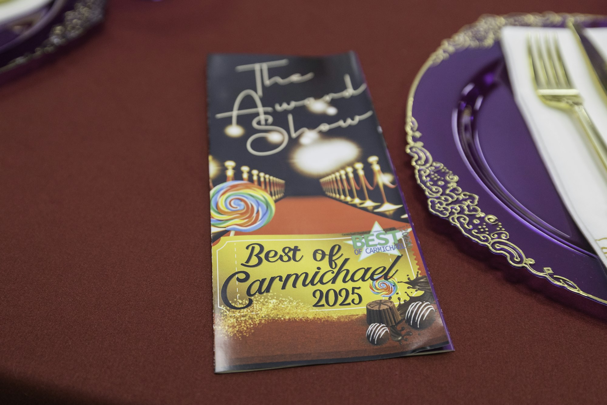 A program labeled "The Award Show: Best of Carmichael 2025" on a red tablecloth beside an ornate purple plate with cutlery.