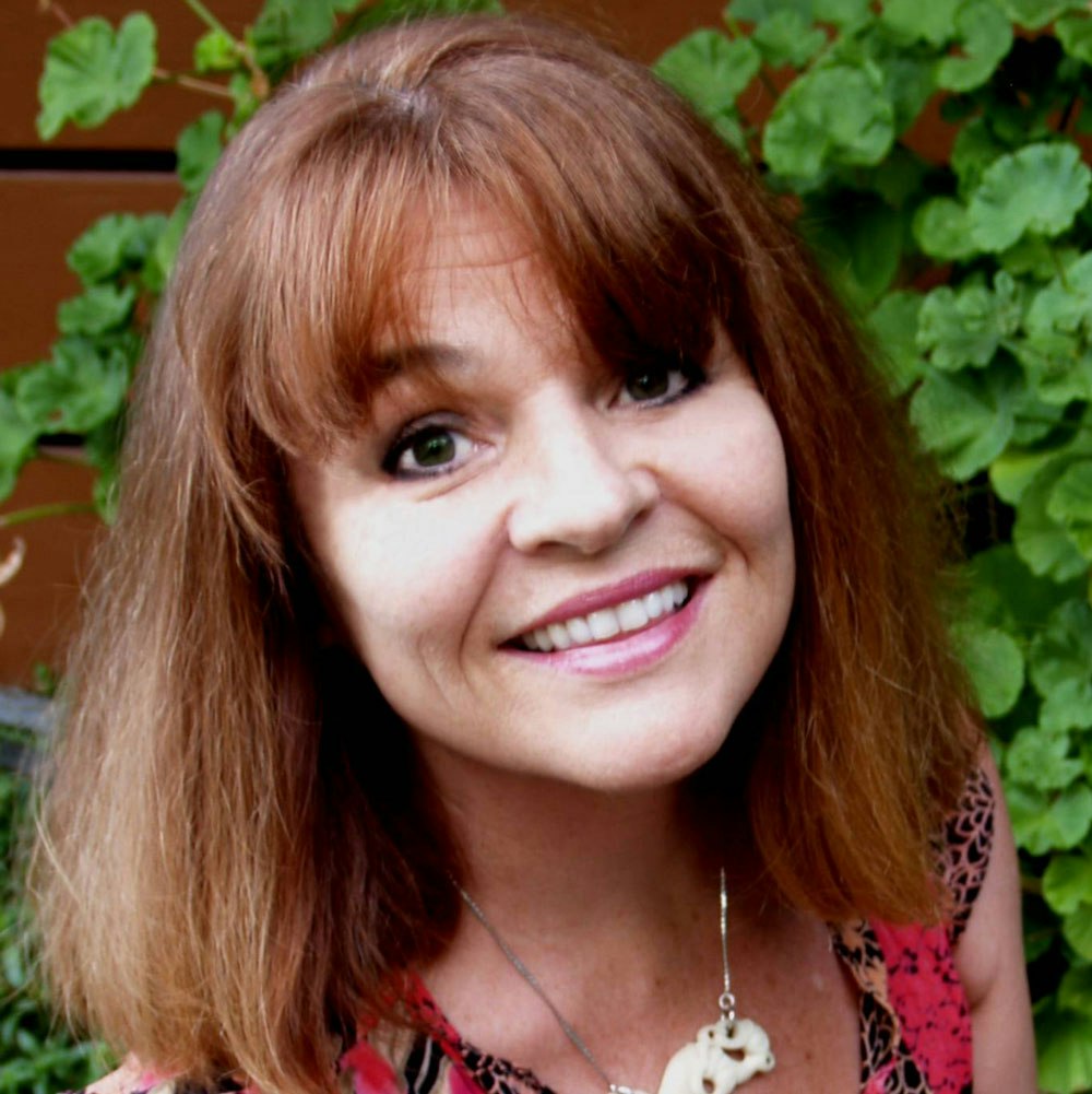 A woman with brown hair smiling, wearing a necklace, in front of green leaves.