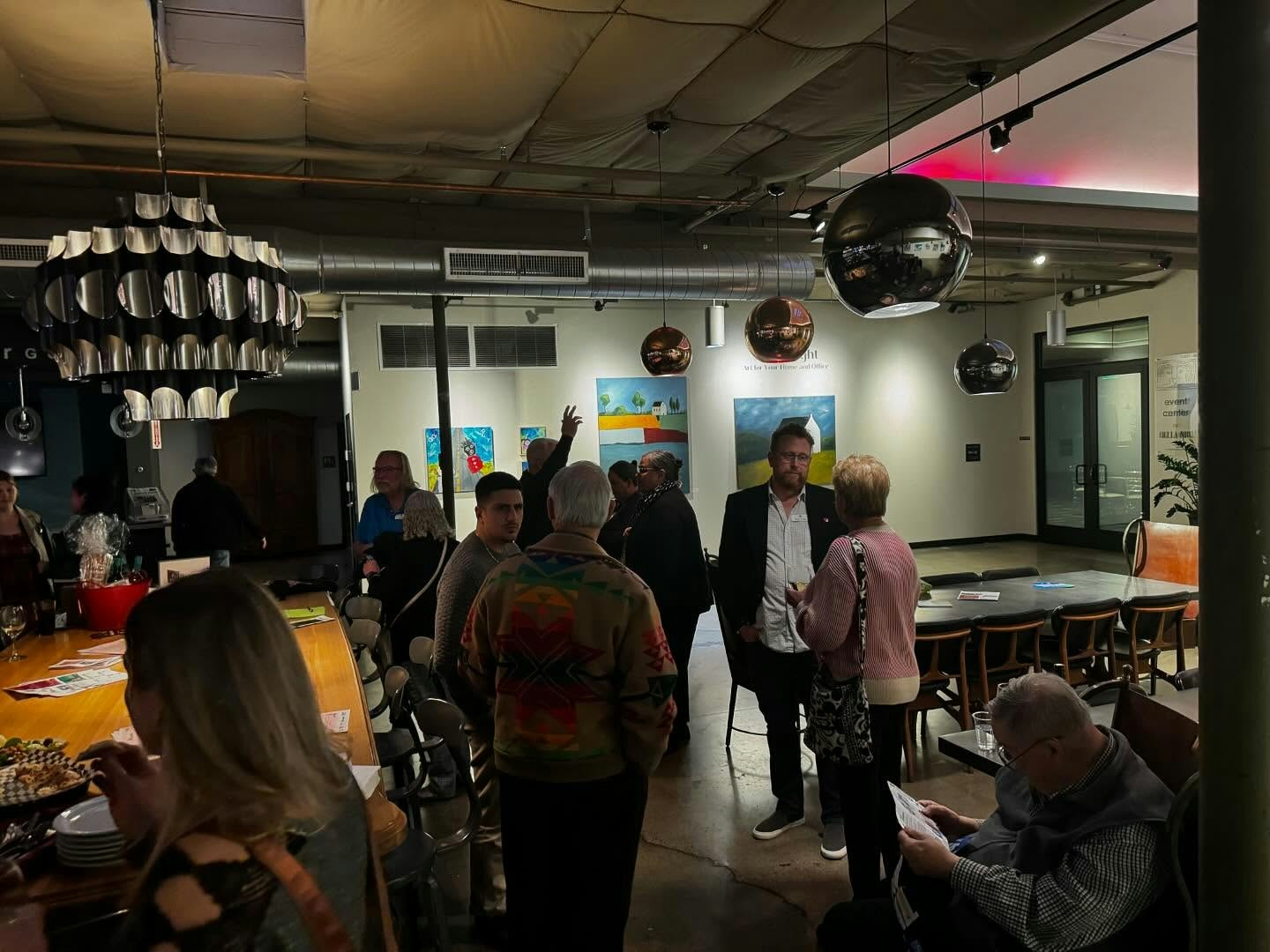 A group of people socializing in a modern space with art on the walls and pendant lights hanging from the ceiling.