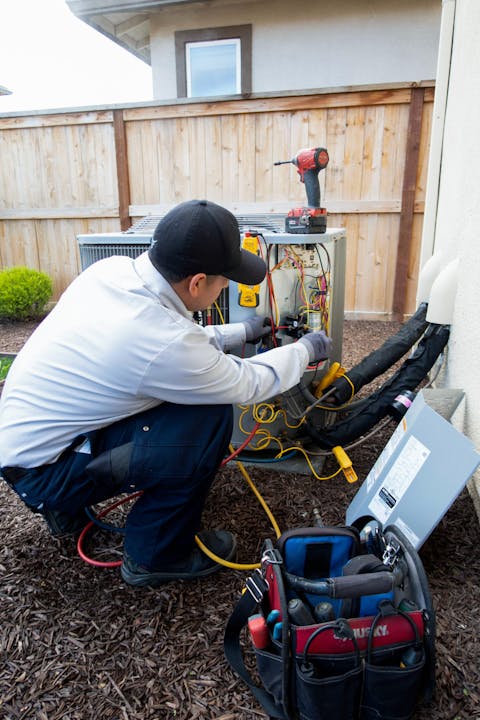 A technician is repairing an outdoor air conditioning unit, surrounded by tools and equipment, next to a wooden fence.