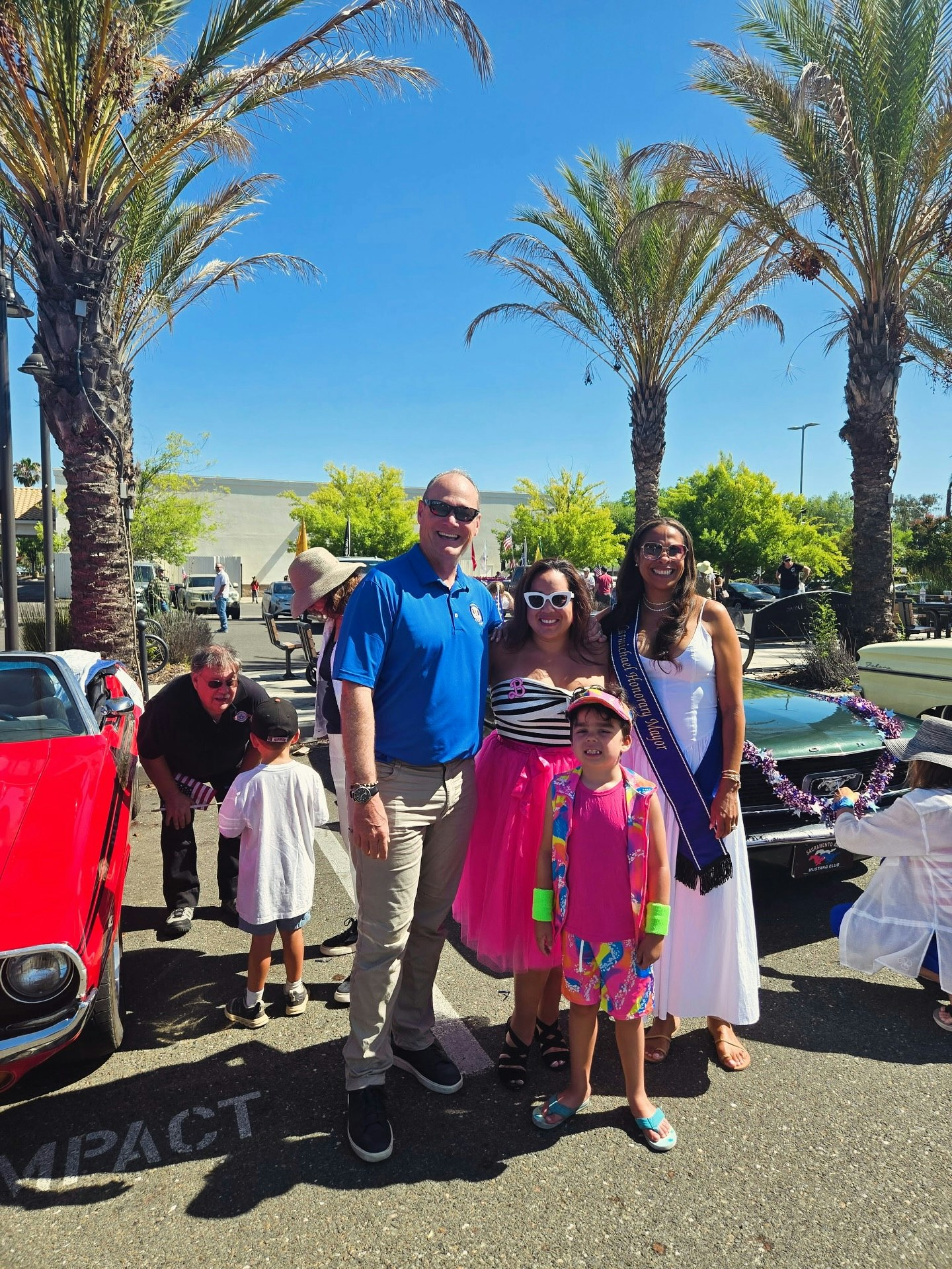 A group of people poses between two colorful cars on a sunny day. Palm trees and blue skies are in the background.