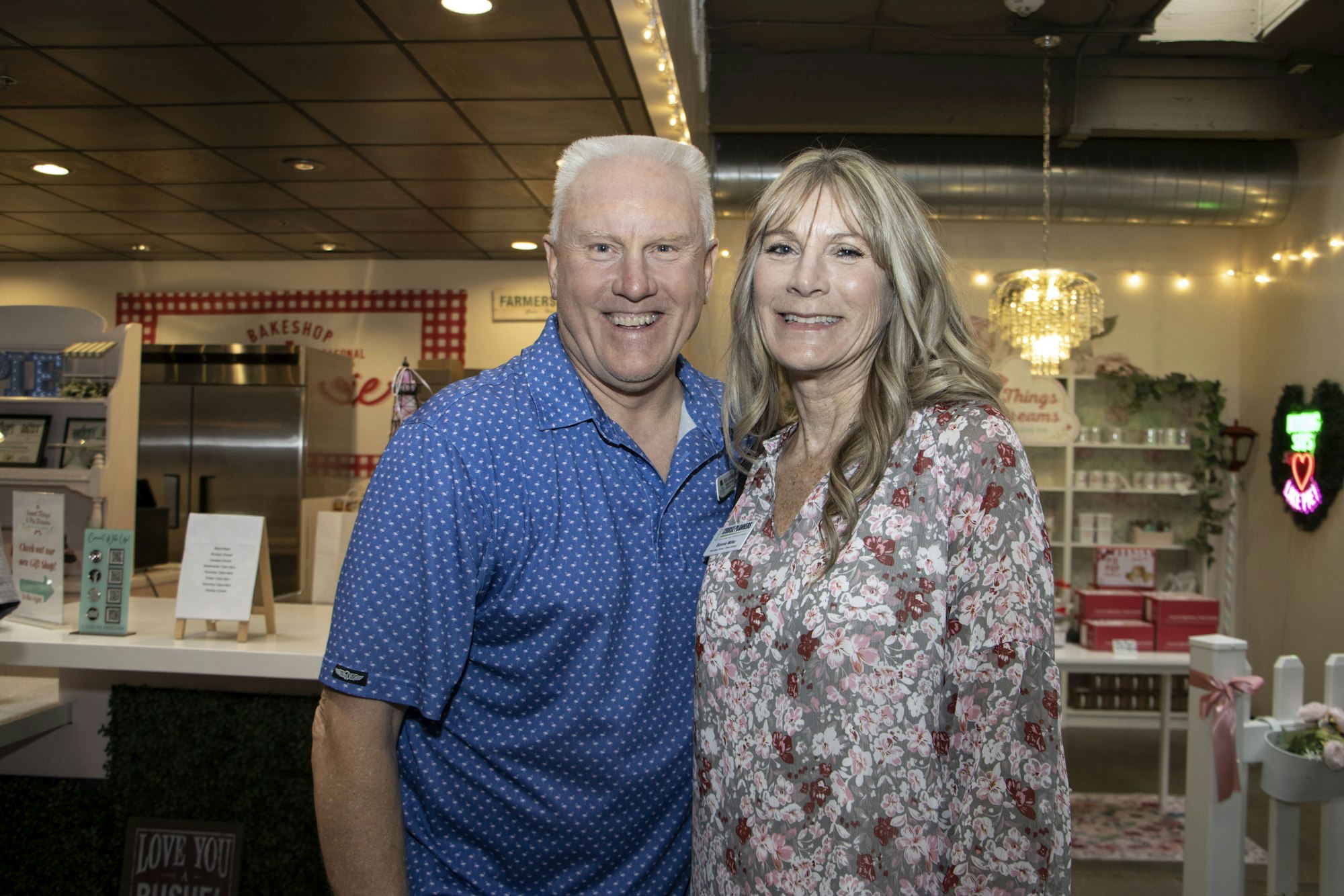 A smiling couple stands together in a cozy, decorated indoor setting with lights and floral patterns.