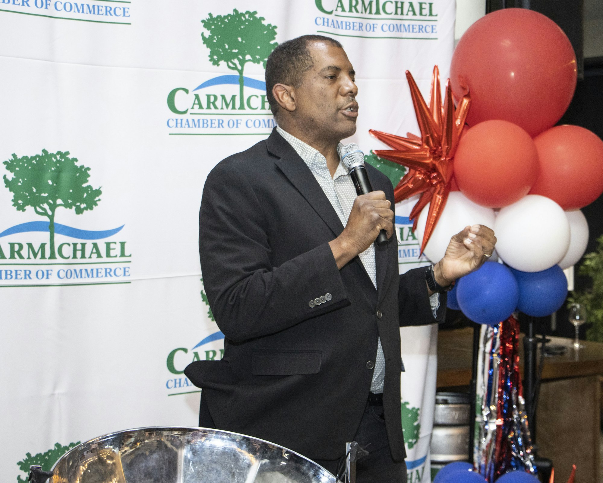 A man speaks at a Carmichael Chamber of Commerce event, with a mic and red, white, and blue balloons in the background.