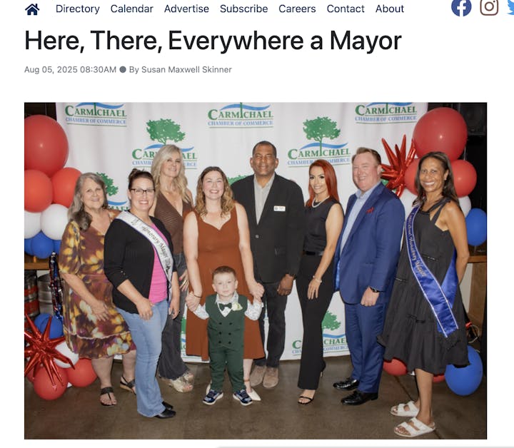 A group of nine people, including a child, pose in front of a "Carmichael Chamber of Commerce" backdrop with festive balloons.