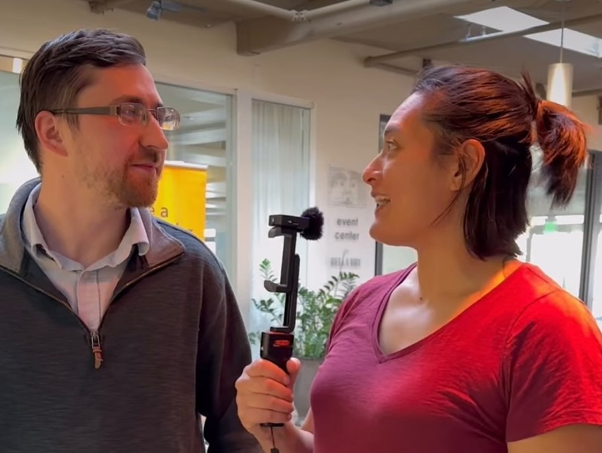 Two people indoors, one with a microphone, seem to be having a conversation or interview in a casual setting.