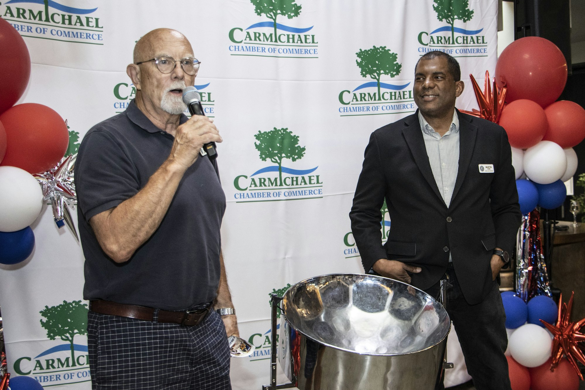 Two men standing in front of a "Carmichael Chamber of Commerce" backdrop with balloons and a steel drum.