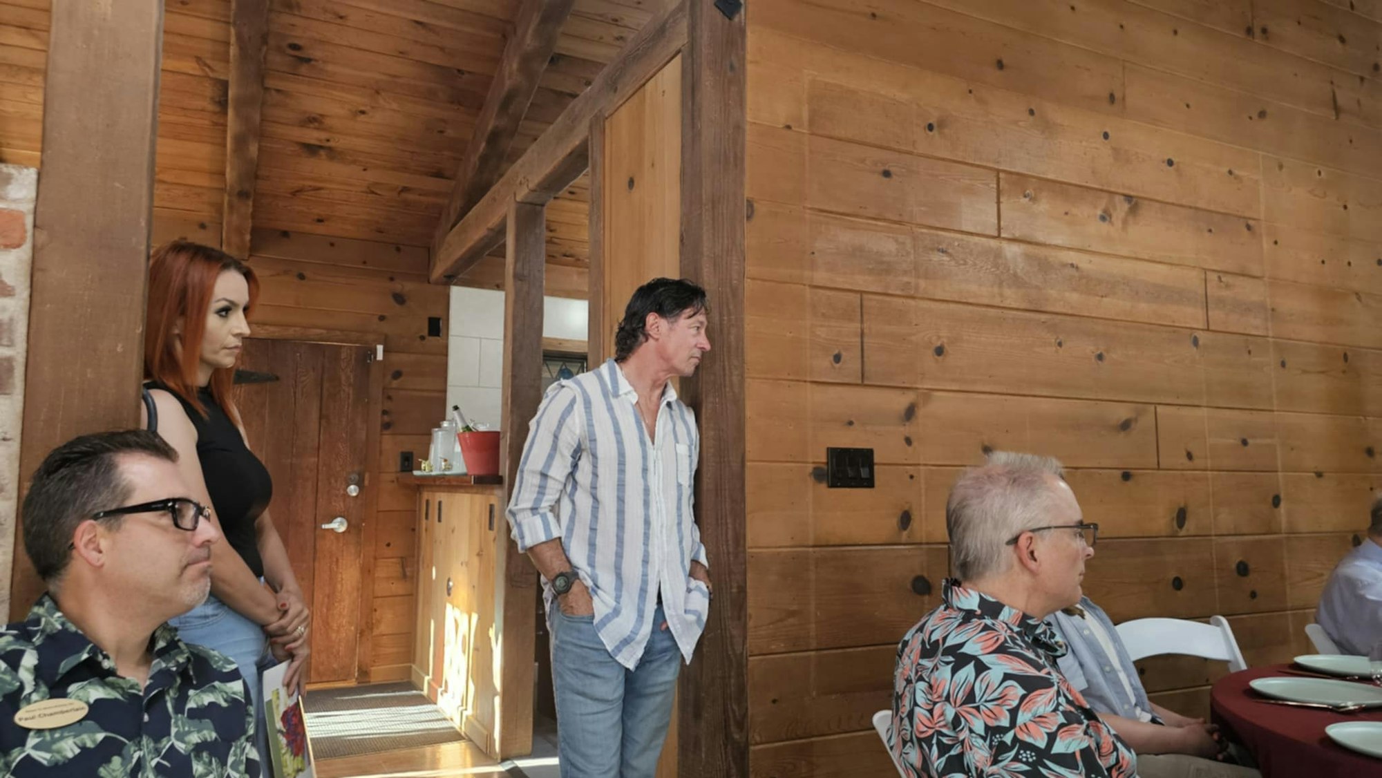 People gathered in a cozy, wooden-walled room, some seated and others standing, with sunlight streaming in.