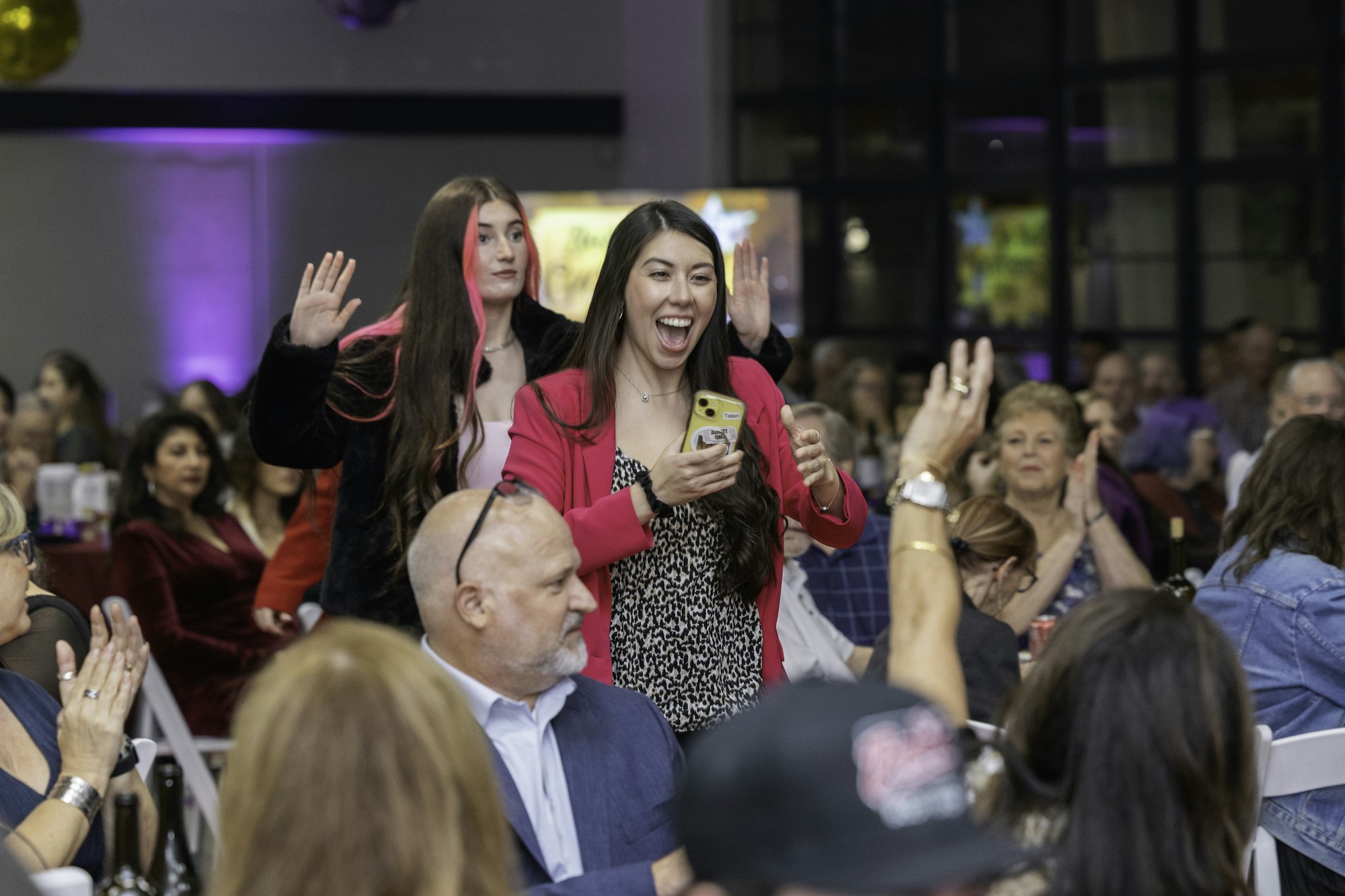 People at a lively event, two women in focus, one in pink, looking excited, others clapping and smiling in the background.
