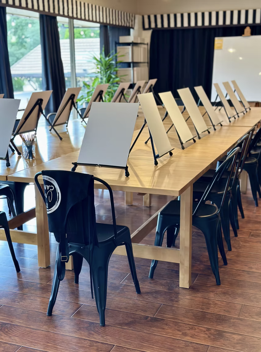 Room with a long table, art easels, chairs, a whiteboard, and natural light.