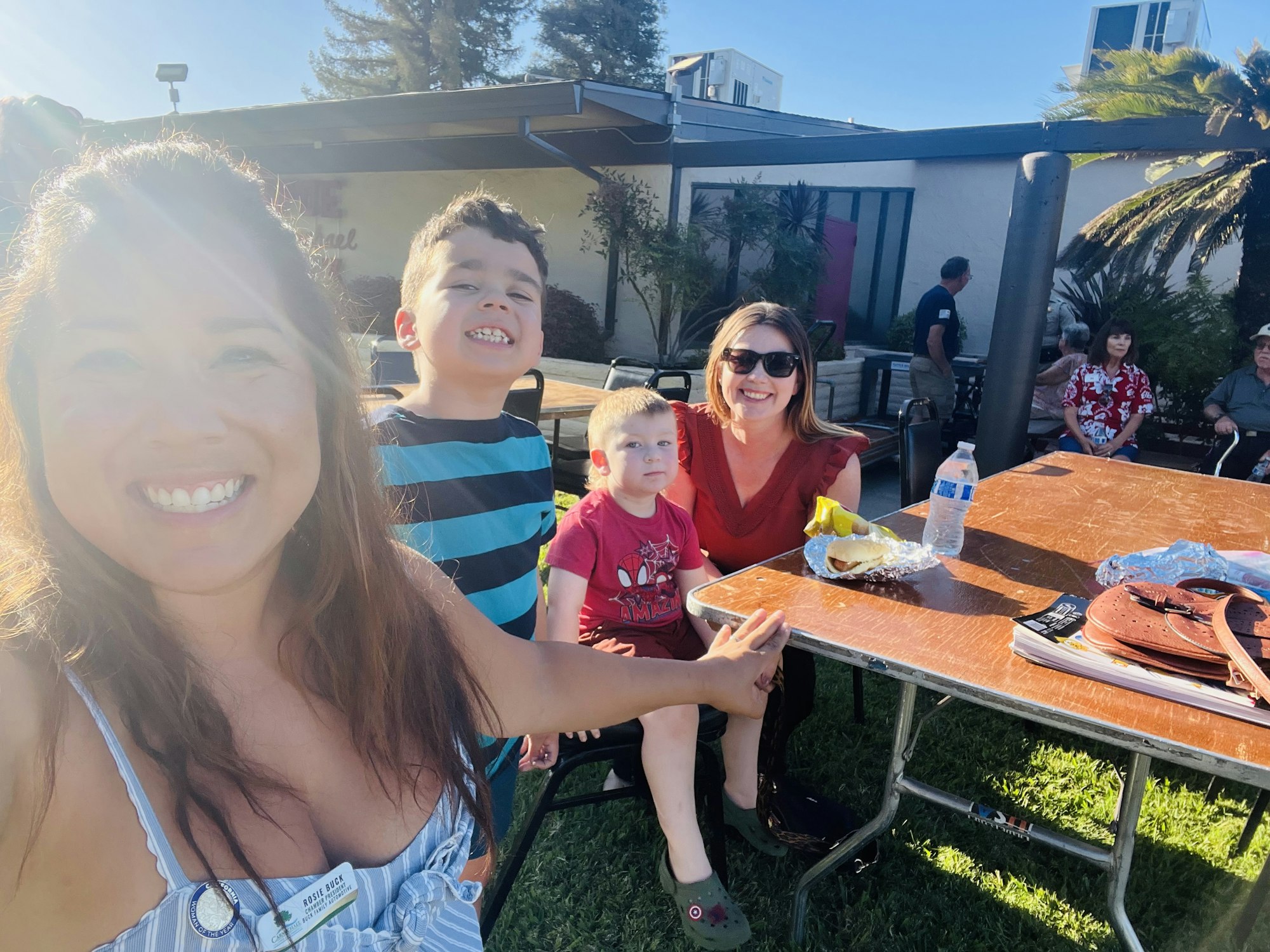 A group of people smiling outdoors at a table with food and a handbag.