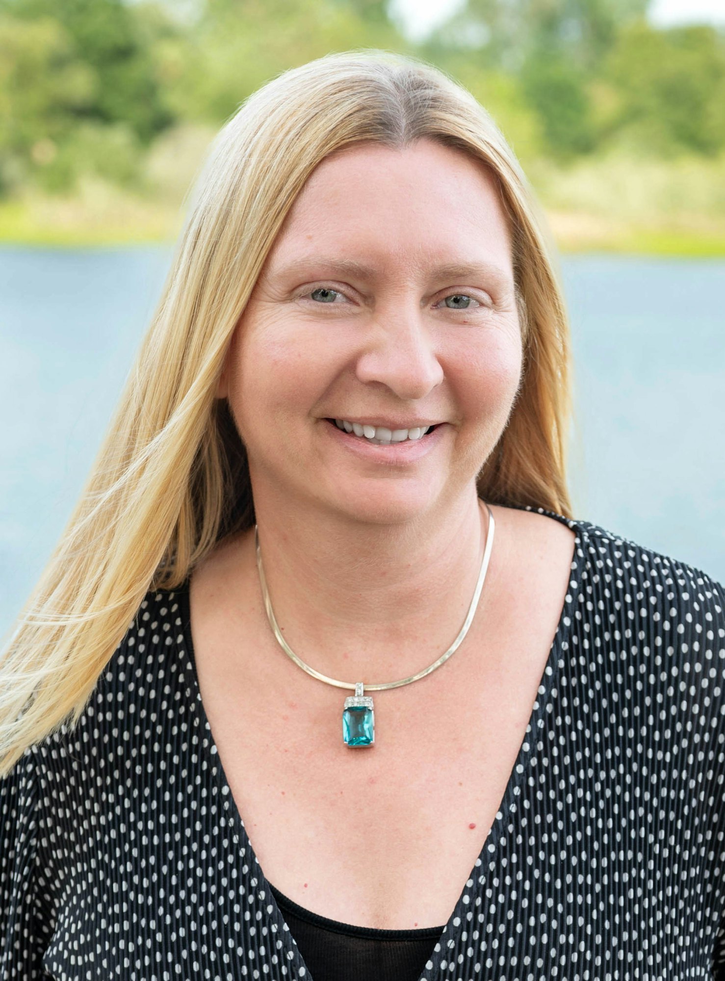 A person smiling outdoors, wearing a polka dot top and a necklace with a blue pendant.