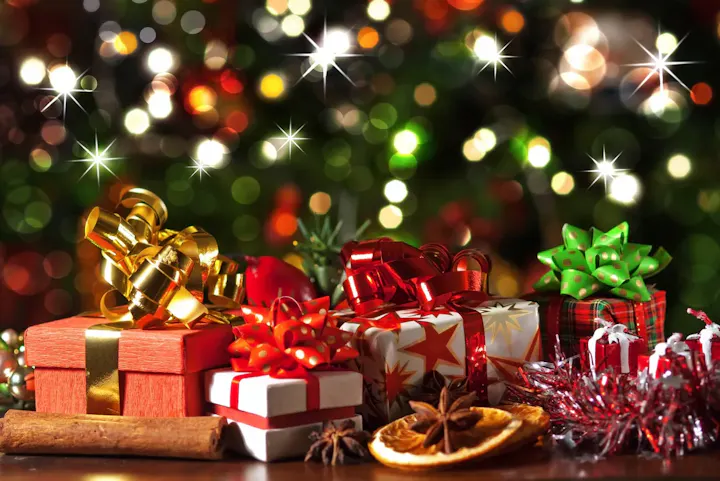 The image features a festive arrangement of colorful wrapped Christmas gifts, ribbons, and seasonal decorations against a blurred background.