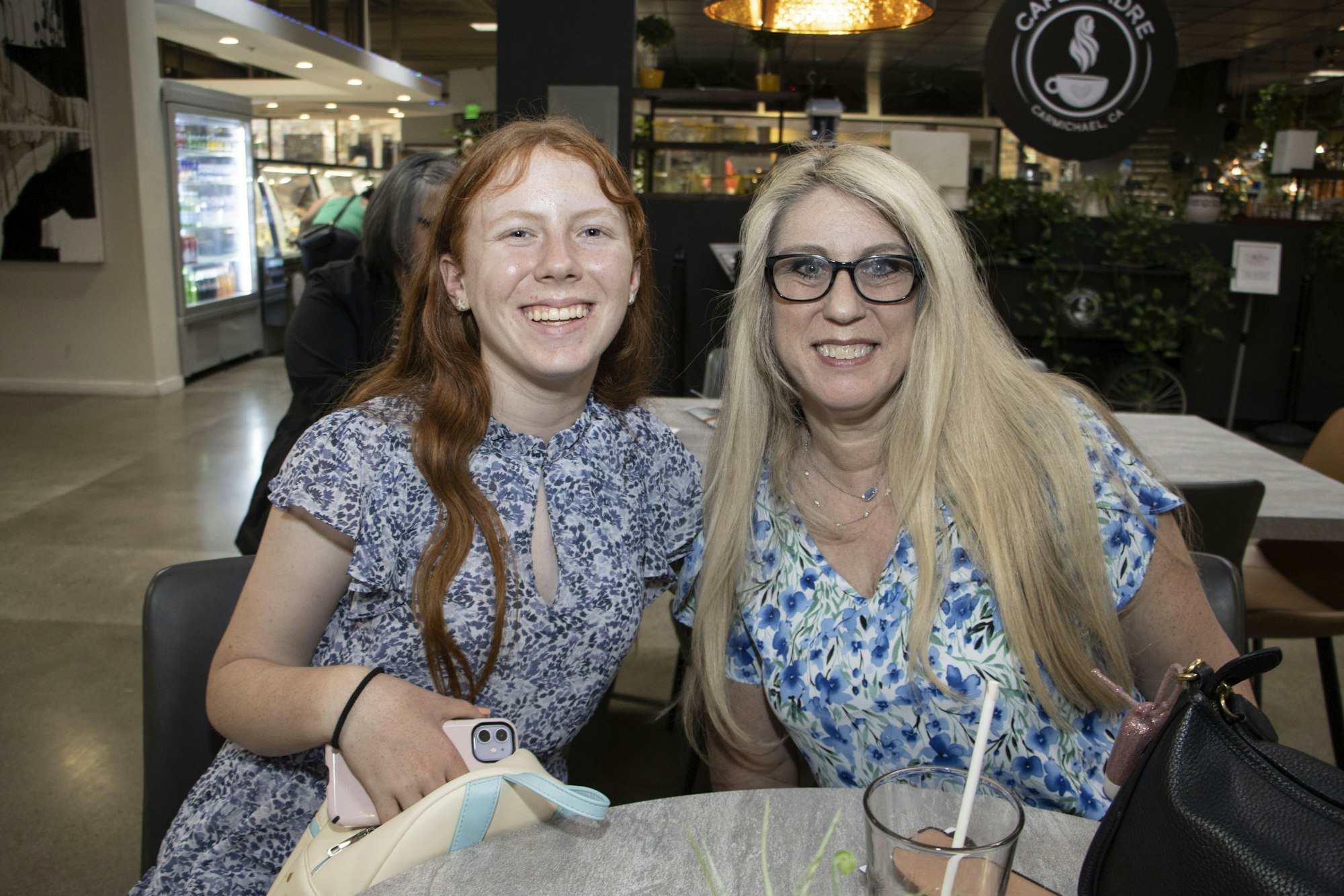 Two people smiling at a cafe table, holding phones, with beverages nearby.
