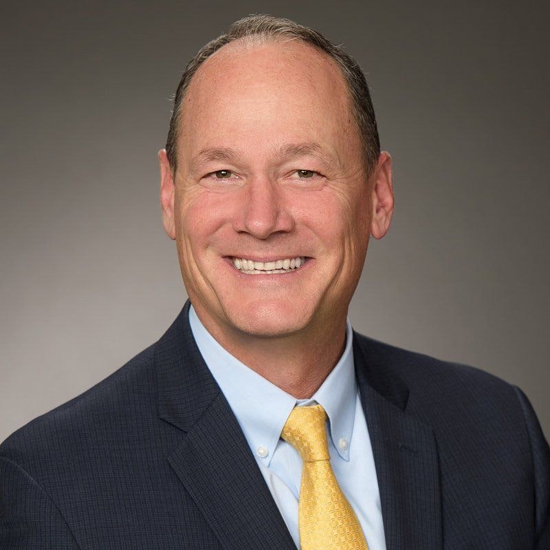 A smiling man in a suit with a blue jacket, light blue shirt, and yellow tie.