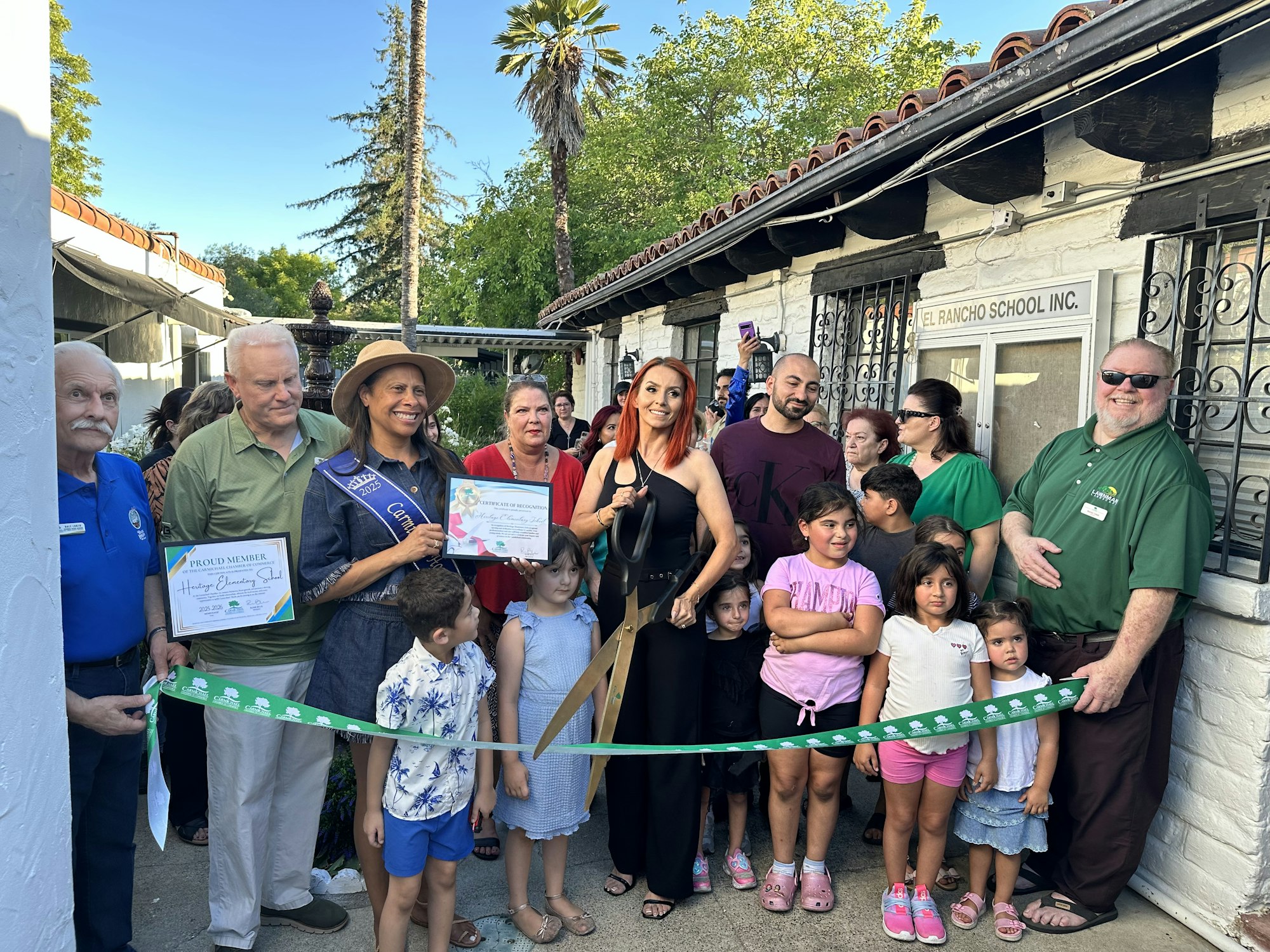 A group of people at a ribbon-cutting ceremony outside "El Rancho School Inc." with certificates and large scissors.