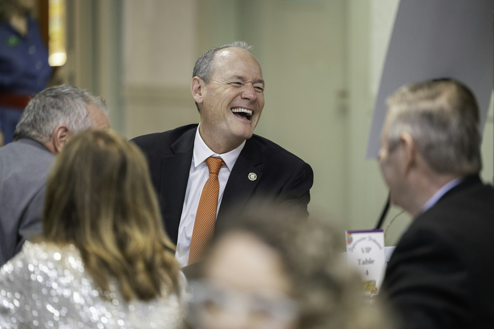 A group of people in formal attire, with one man smiling and laughing, sitting at a table with a "VIP Table 4" sign.