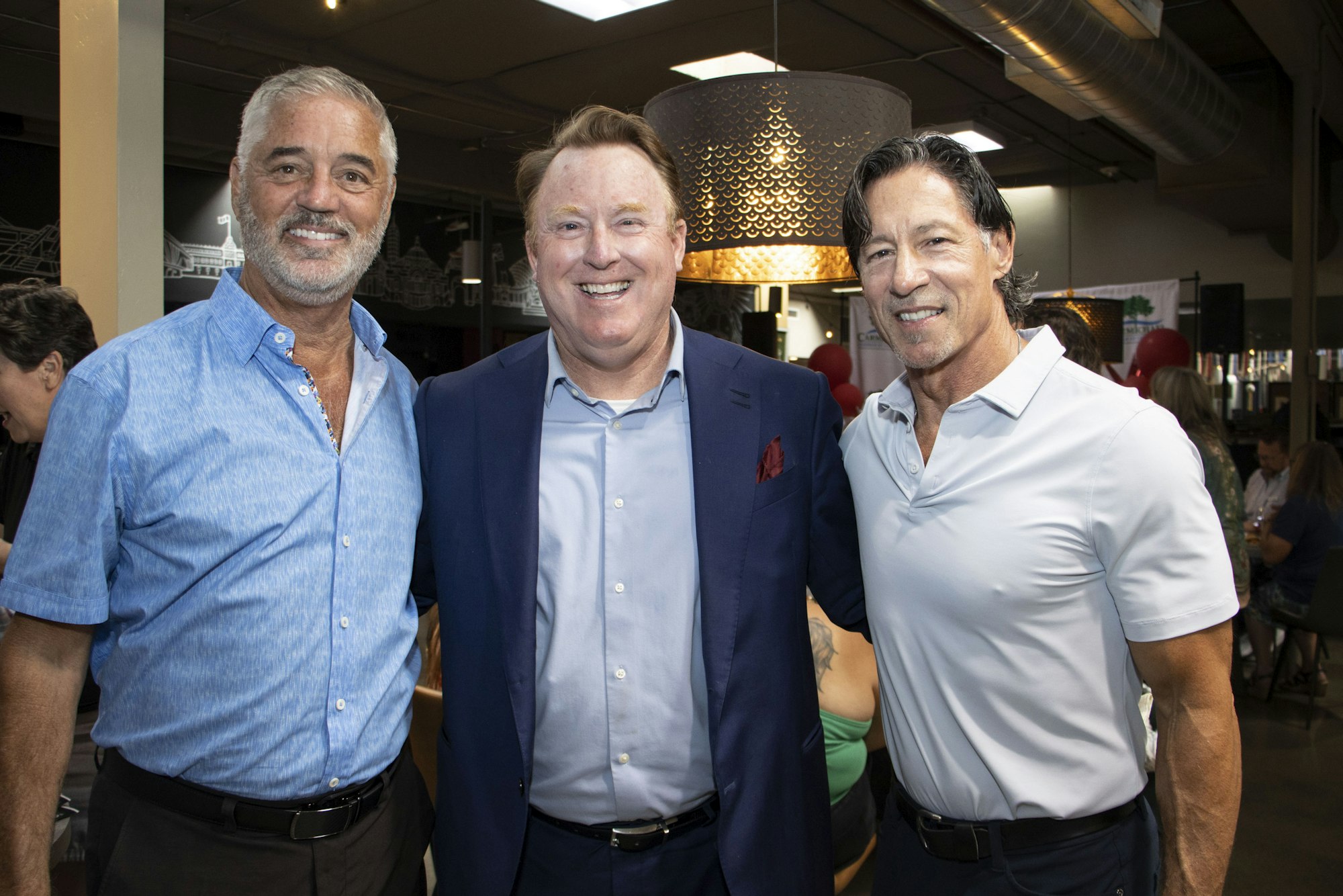 Three men smiling, posing together indoors, two in casual shirts, one in a suit.