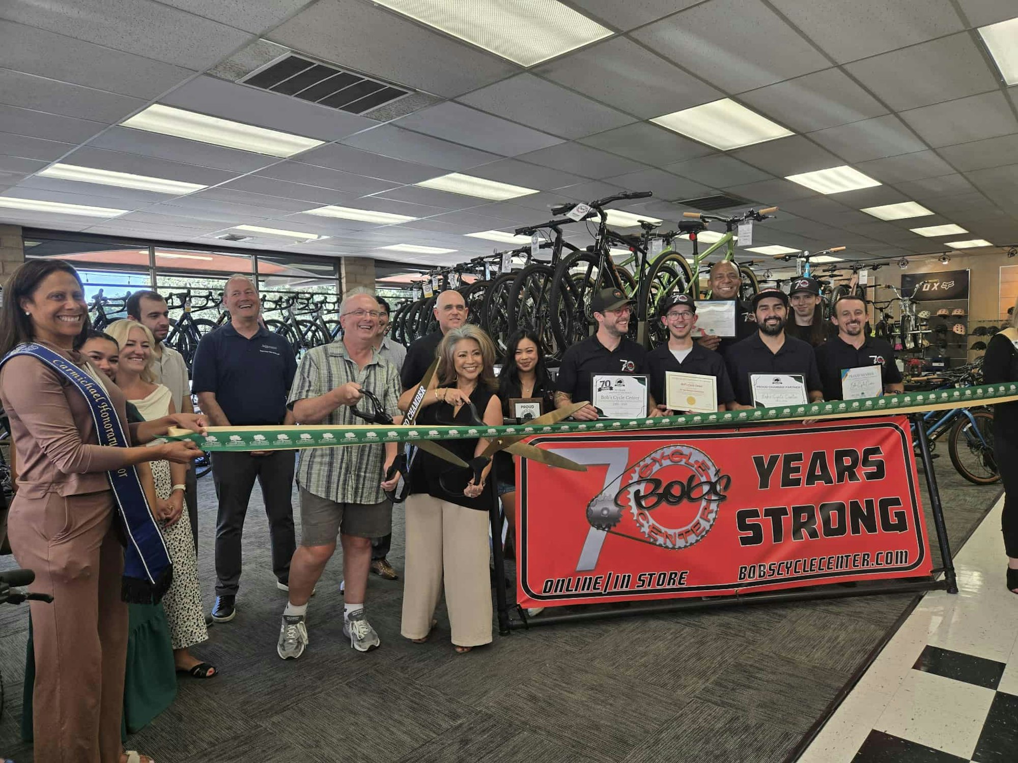 A group celebrating a ribbon-cutting ceremony for a bike shop's 70th anniversary, with bikes in the background.