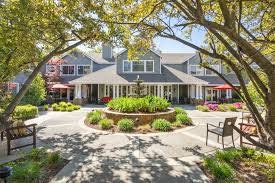 A charming house with a landscaped front yard, featuring a round flower bed and patio seating under shady trees.