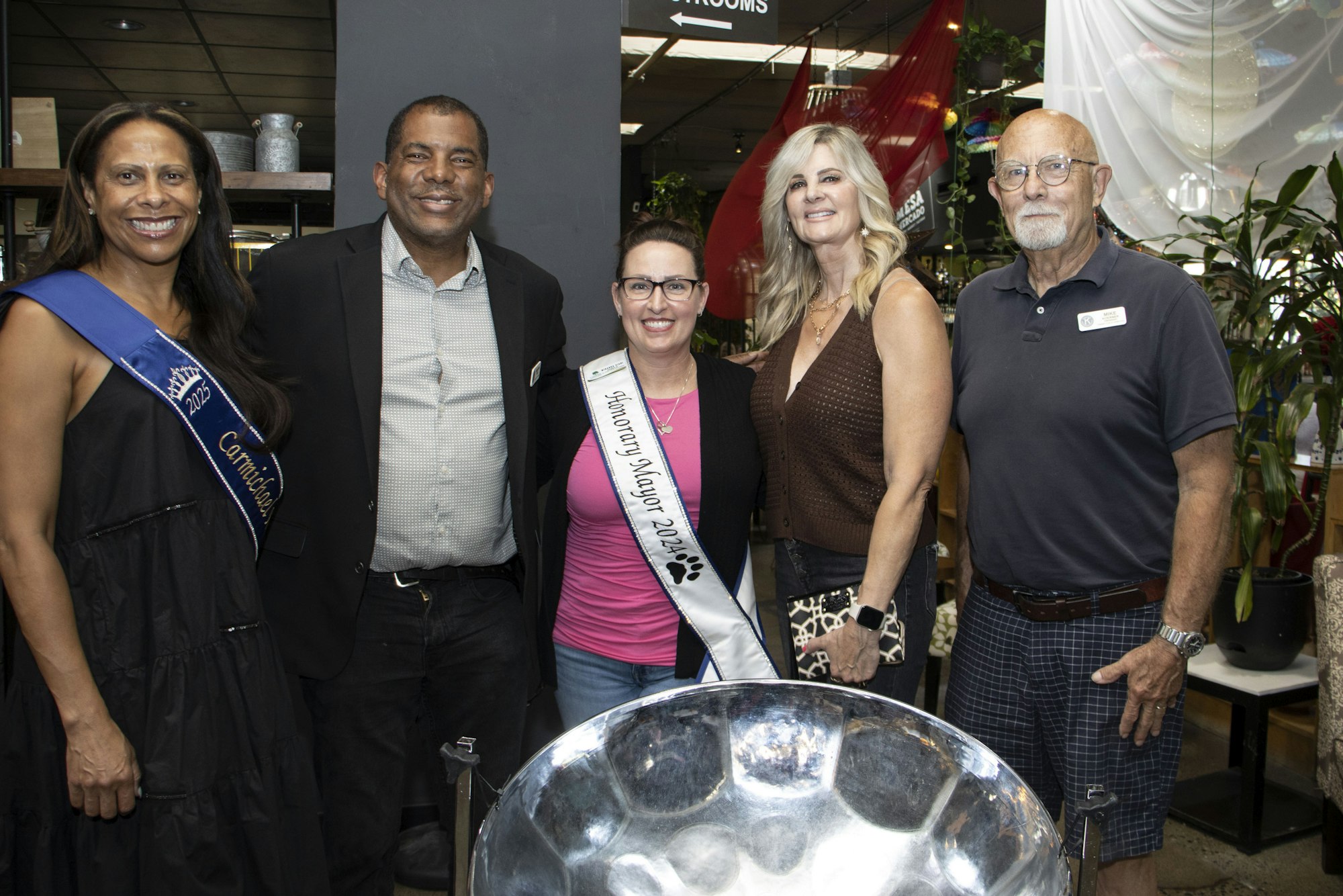 Five people posing, two wearing sashes with text. Indoors setting with plants and decorations visible in the background.