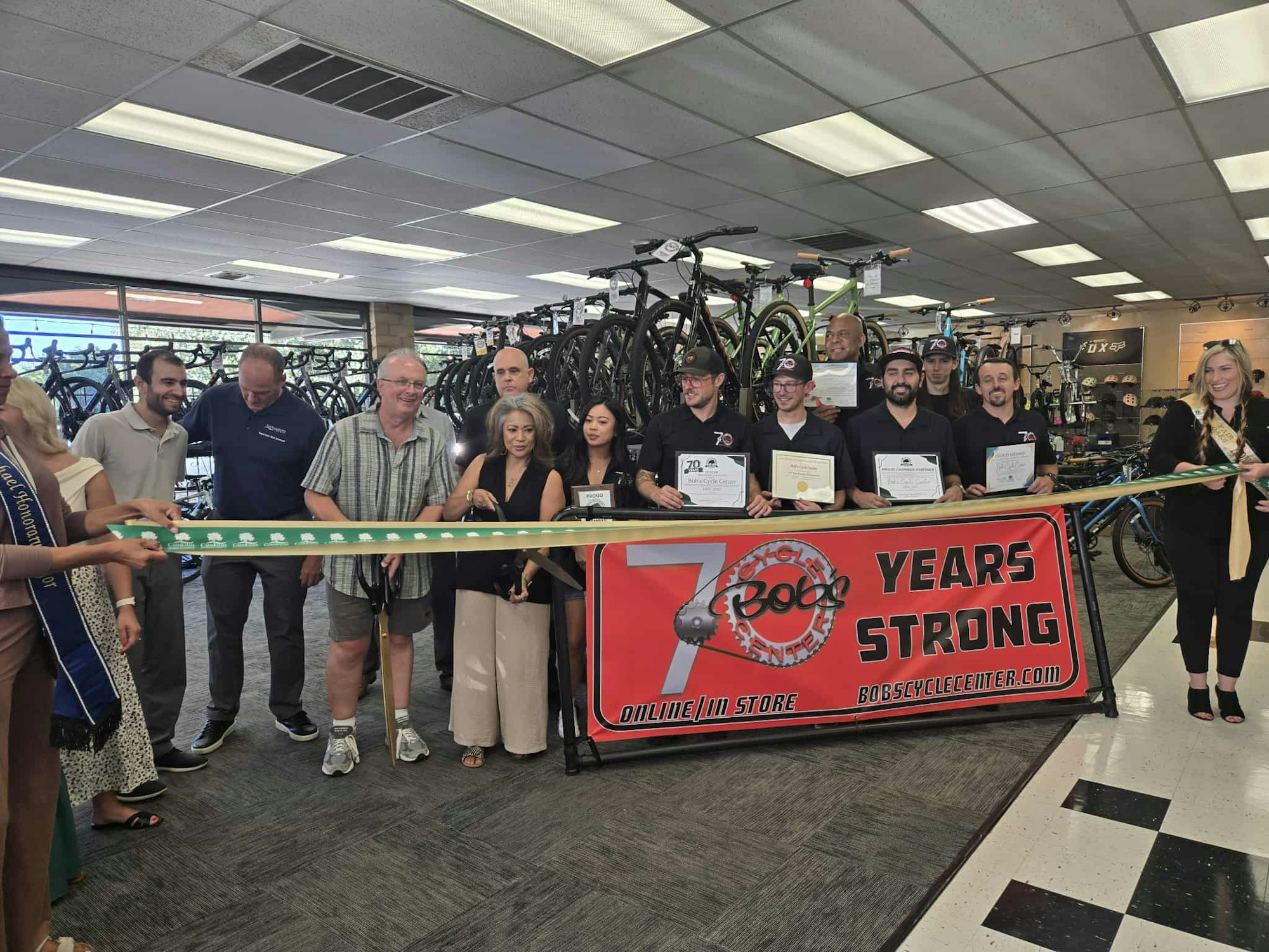 People are at a ribbon-cutting event in a bike shop celebrating 70 years. They hold a banner and some are holding certificates.