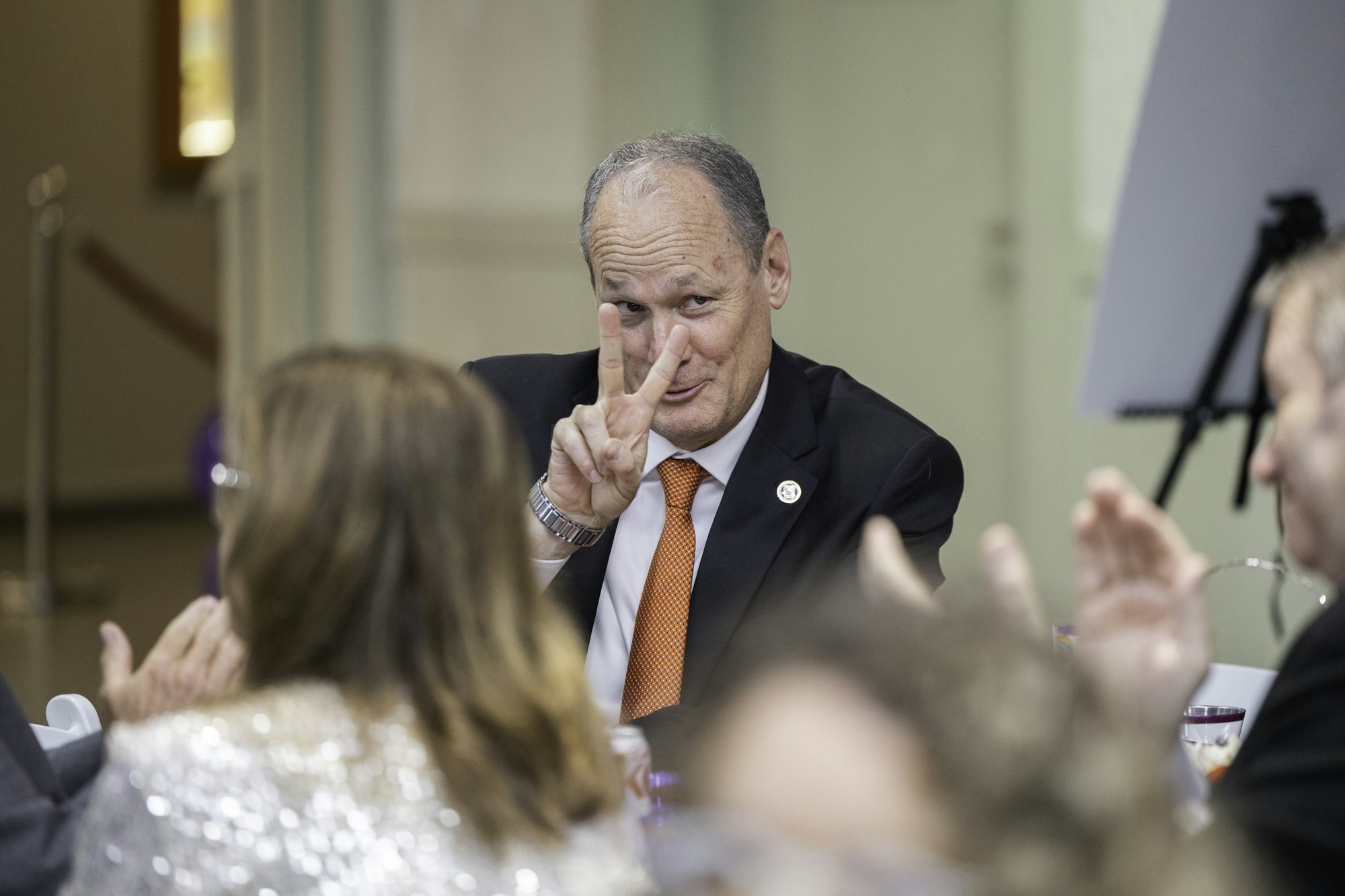 A man in a suit makes a peace sign with his fingers at a seated event.
