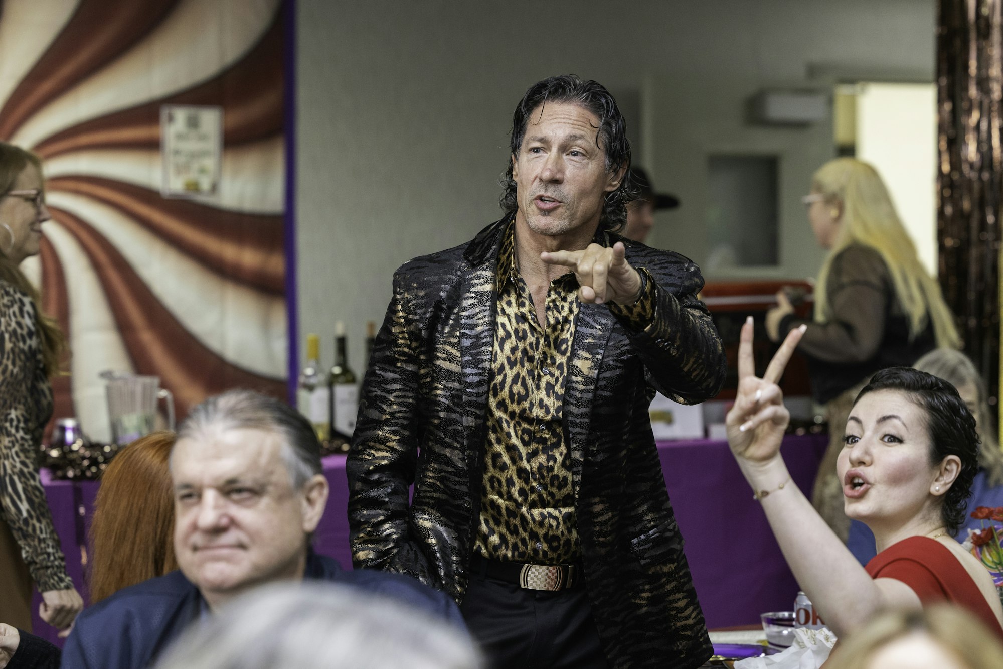 A man in a patterned outfit gestures while a woman nearby raises two fingers in a peace sign, at a lively gathering.
