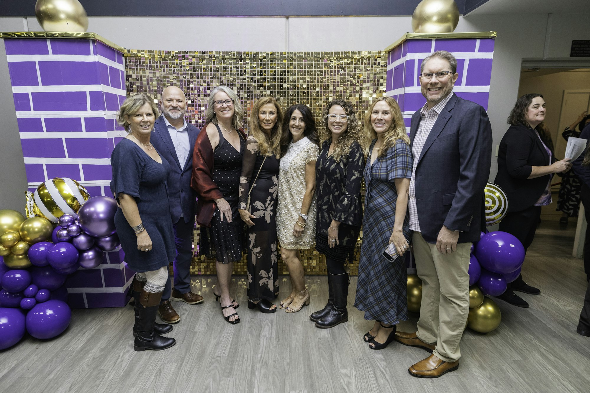 A group of people posing in front of a sequin backdrop with purple and gold balloon decorations.