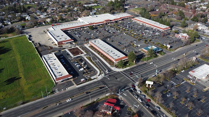 Aerial view of a shopping center with parking lots, roads, and surrounding greenery.