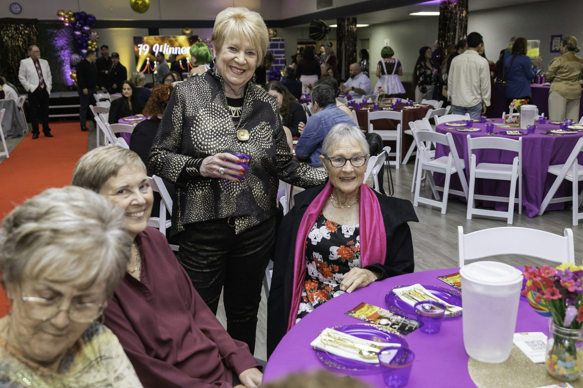 People at a festive event with purple tablecloths and red carpet. A woman stands smiling beside seated guests.