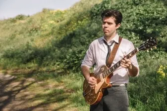 A man in a white shirt and tie stands outdoors playing a guitar, surrounded by greenery and sunlight.