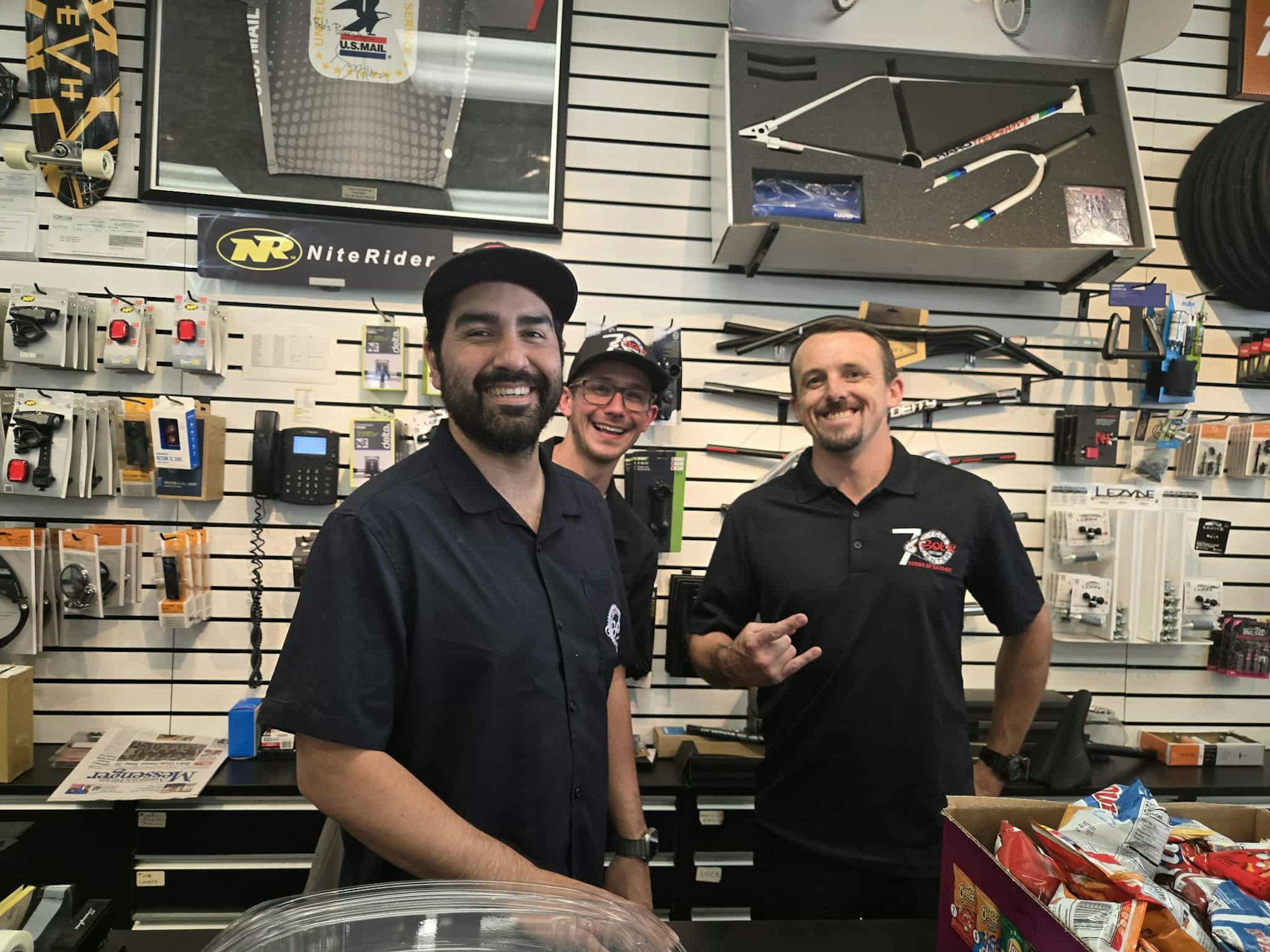 Three people smiling in a store with various biking accessories displayed on the wall.