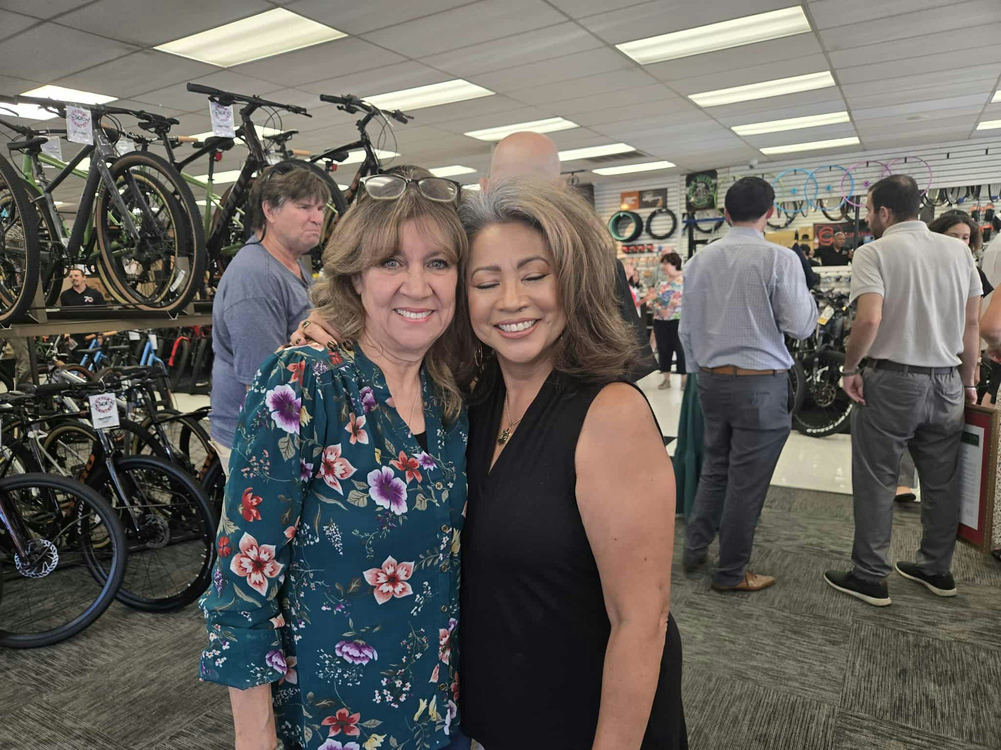 Two women smiling and hugging in a bike shop with people and bicycles in the background.