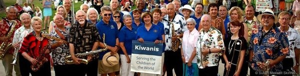 Group of smiling people holding a "Kiwanis" sign, some with musical instruments, outdoors in casual attire.