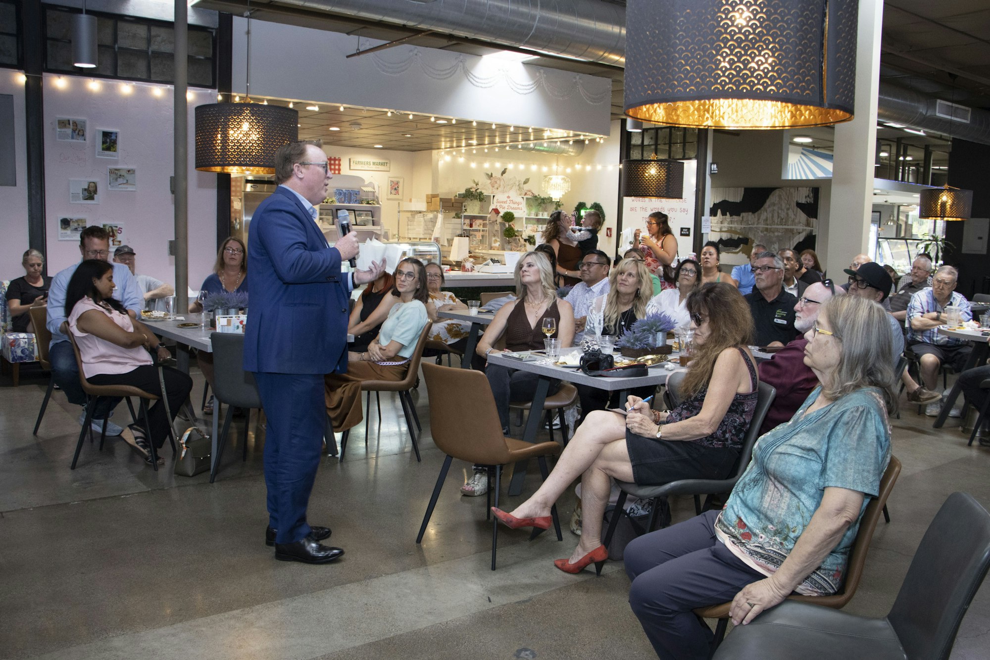 A man in a blue suit speaks to an attentive audience seated at tables in a modern, well-lit restaurant or cafe.