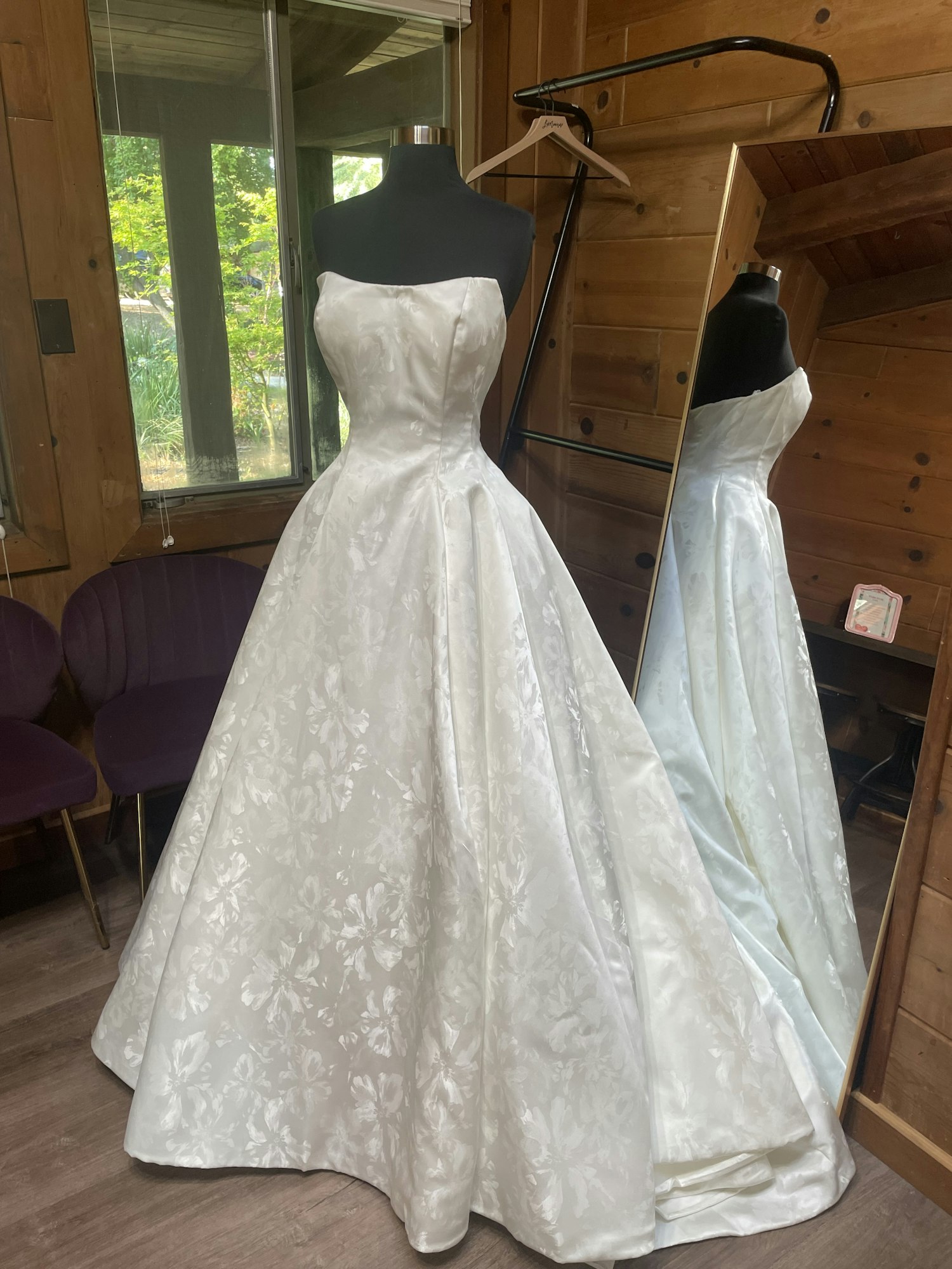 A white strapless dress with floral patterns on a mannequin in a wood-paneled room, reflected in a mirror.