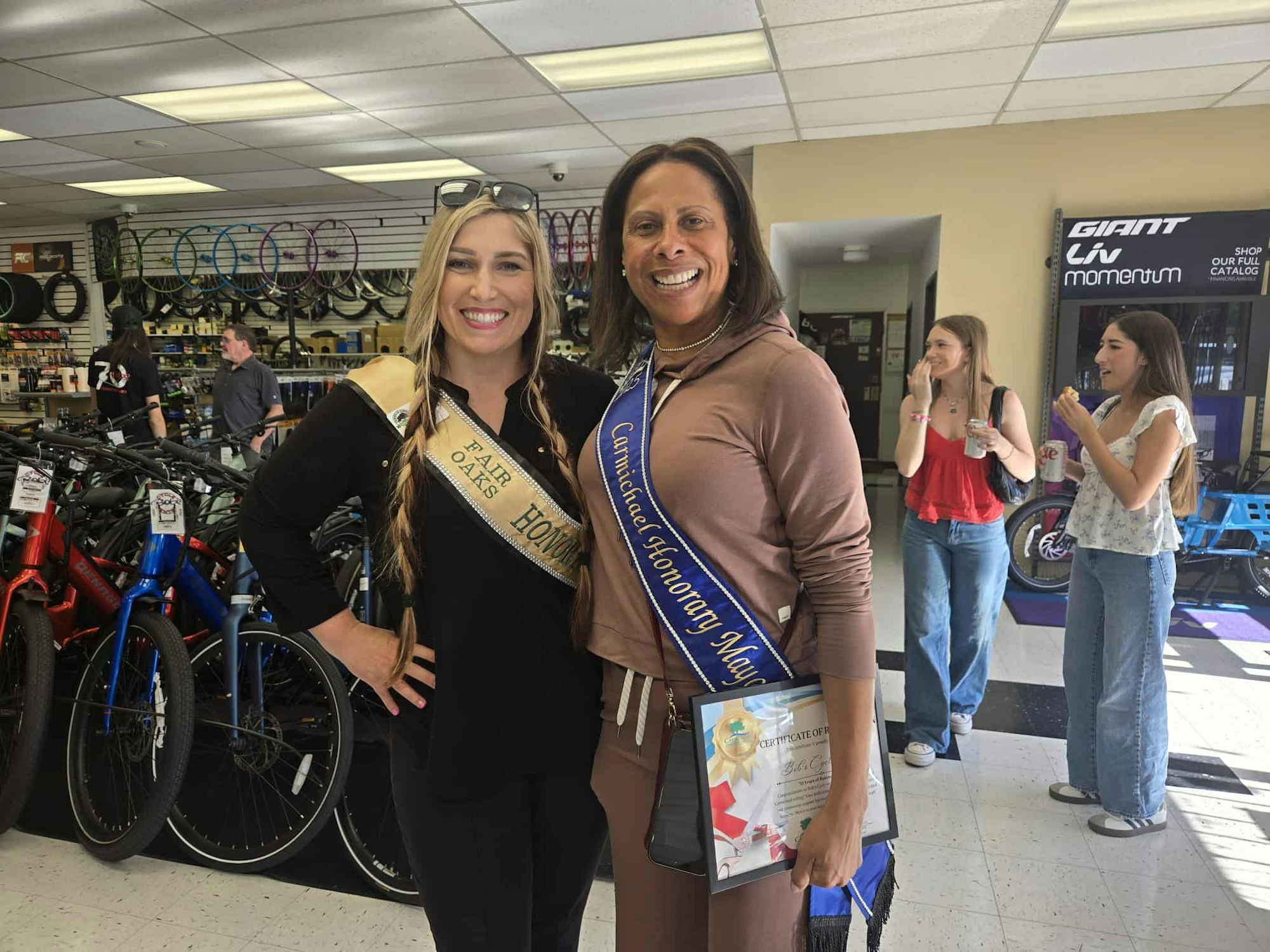 Two women wearing sashes are smiling in a bike shop. One holds a certificate. Two people are in the background.