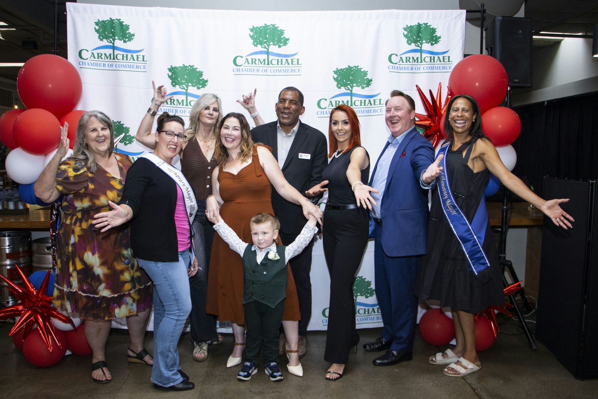 A group of smiling people pose in front of a "Carmichael Chamber of Commerce" banner with red, white, and blue balloons.