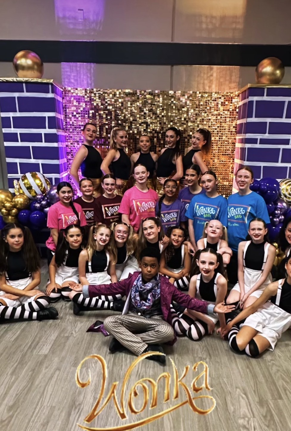A group of people dressed in colorful outfits with a "Wonka" theme, posing for a photo with a decorative backdrop.