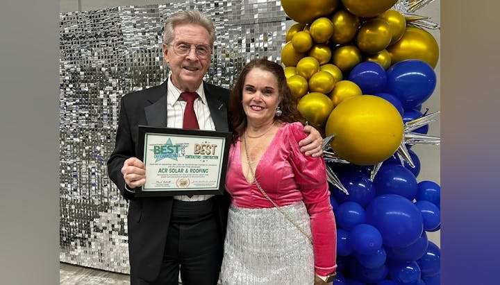Two people smiling, holding an award certificate, with a backdrop of silver tinsel and yellow and blue balloons.