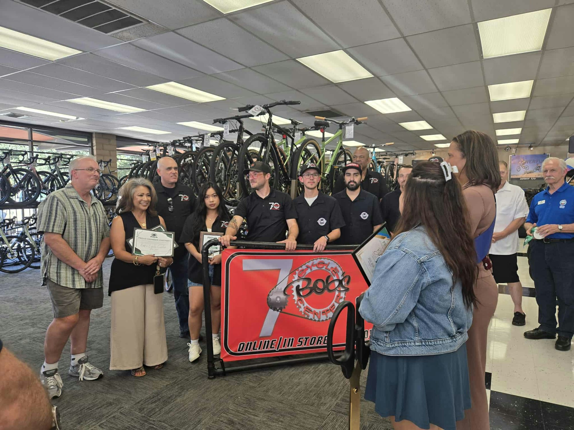 A group of people at a bicycle shop event, some holding certificates, with bikes displayed in the background.