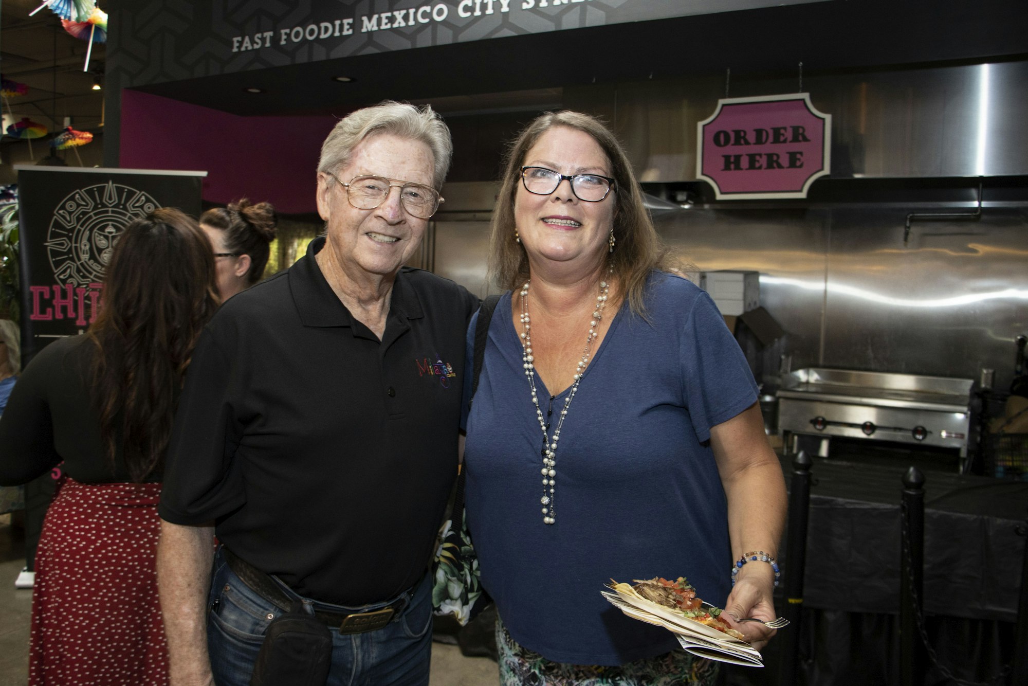 Two people smiling at a restaurant with a sign that says "ORDER HERE" and "FAST FOODIE MEXICO CITY STYLE."