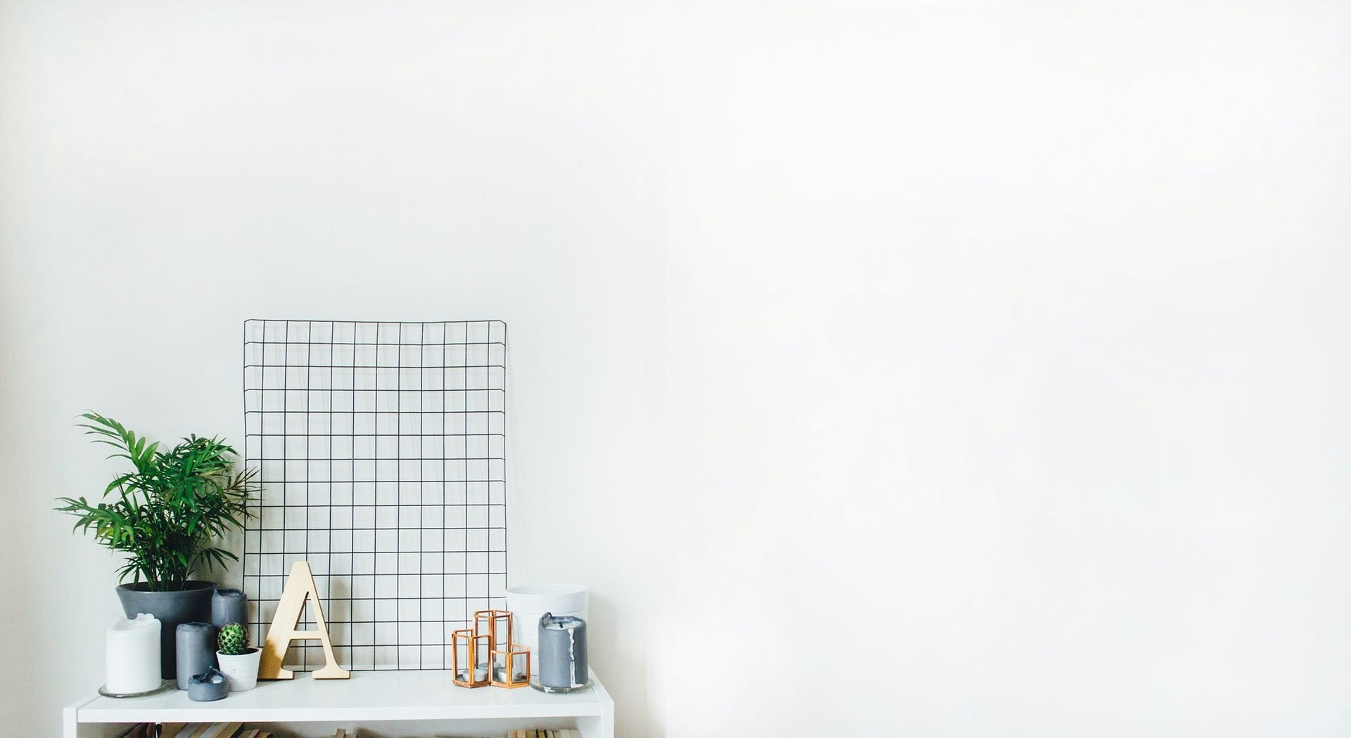Minimalist shelf decor with plant, candles, geometric objects, and metal grid against a white wall.