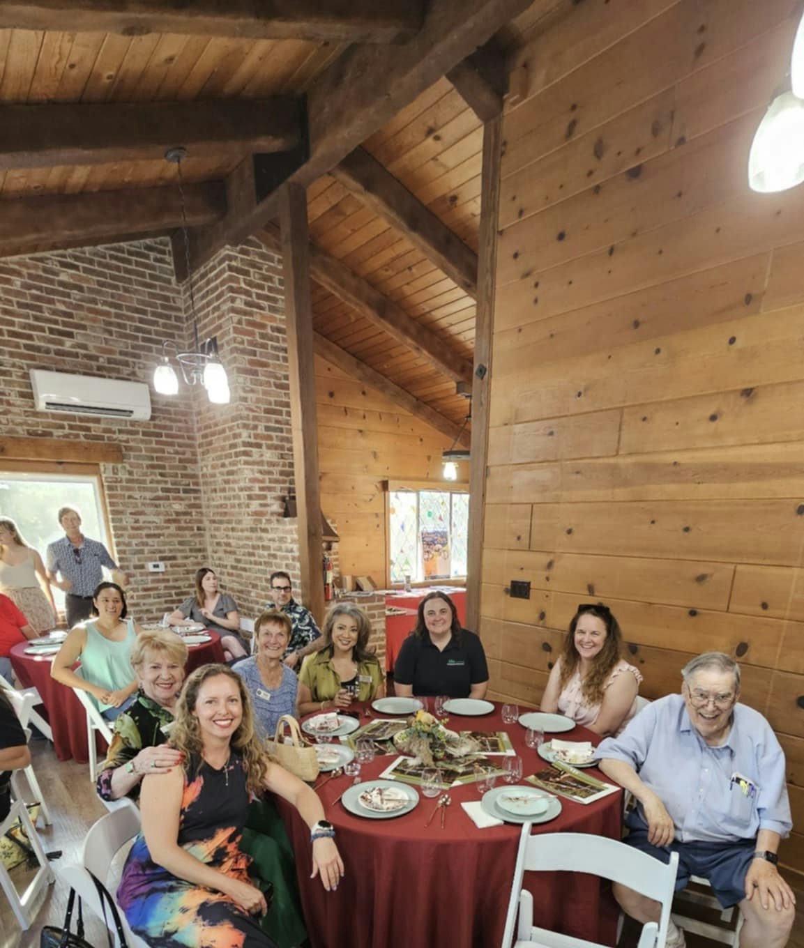 A group of people are dining in a cozy, wooden interior with a brick wall, seated around tables with red tablecloths.
