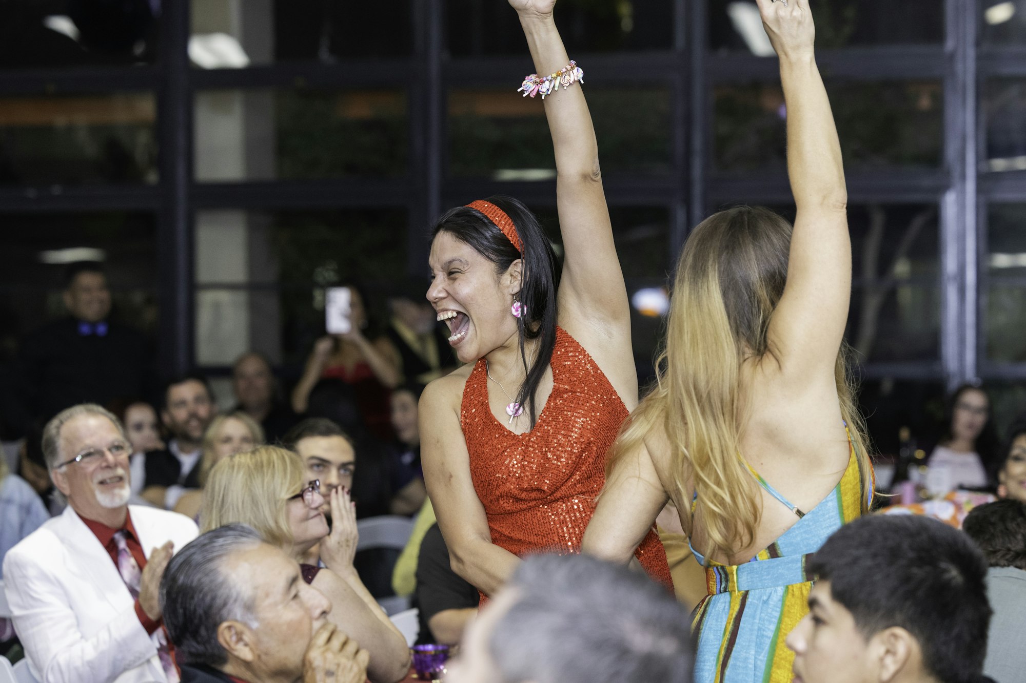 A woman in a red dress is celebrating with people clapping and cheering around her at an indoor event.