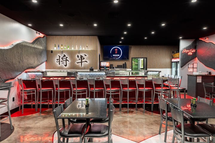 A sushi bar with red chairs, modern decor, and a "Shogun" sign above a counter with bottles and a TV screen.