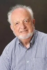 The image features an older man with gray hair and a beard, wearing a checkered shirt, smiling against a plain background.