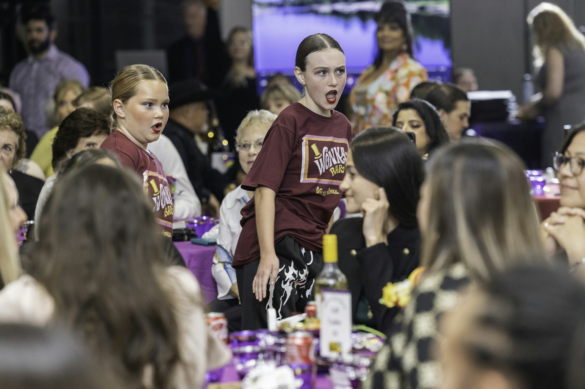 Two children in "Wonka" shirts performing at an event with seated audience.